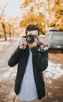A person is capturing a photograph on a tree-lined street with autumn foliage. The individual is wearing a dark quilted jacket over a striped shirt, positioning a camera up to their face while focusing on the scene ahead.