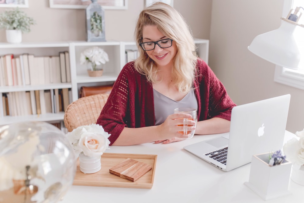 MacBookを持ってテーブルの横に座っているガラスのマグカップを持って微笑む女性