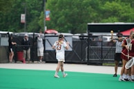 A lacrosse game scene with a player wearing number 23 walking on a green field. Nearby, a referee is holding a lacrosse ball in the air. On the right side, other players in maroon uniforms stand together holding lacrosse sticks. In the background, there are people behind a fence and some greenery.