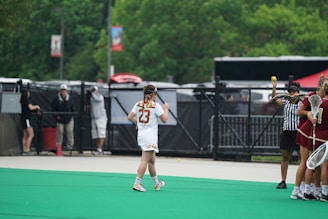 A lacrosse game scene with a player wearing number 23 walking on a green field. Nearby, a referee is holding a lacrosse ball in the air. On the right side, other players in maroon uniforms stand together holding lacrosse sticks. In the background, there are people behind a fence and some greenery.