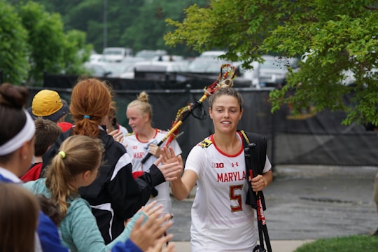 A female lacrosse player in a white jersey with the number 5 is walking and high-fiving a group of excited fans. She carries a lacrosse stick and has her hair in a bun. Other athletes and people are visible in the background, with trees and parked cars lining the area.