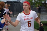 A female athlete in a white jersey with the word 'Maryland' and the number 6, giving high-fives to people on the sidelines. She wears a red headband and appears focused as she walks. Other team members and onlookers are visible in the background.