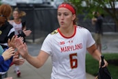 A female athlete in a white jersey with the word 'Maryland' and the number 6, giving high-fives to people on the sidelines. She wears a red headband and appears focused as she walks. Other team members and onlookers are visible in the background.