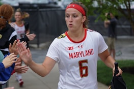 A female athlete in a white jersey with the word 'Maryland' and the number 6, giving high-fives to people on the sidelines. She wears a red headband and appears focused as she walks. Other team members and onlookers are visible in the background.