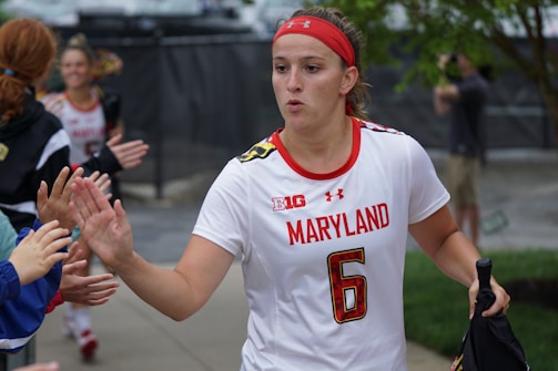 A female athlete in a white jersey with the word 'Maryland' and the number 6, giving high-fives to people on the sidelines. She wears a red headband and appears focused as she walks. Other team members and onlookers are visible in the background.