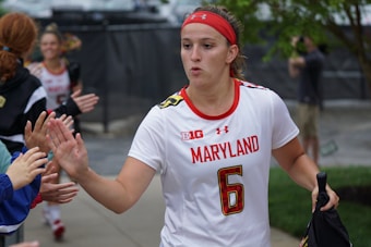 A female athlete in a white jersey with the word 'Maryland' and the number 6, giving high-fives to people on the sidelines. She wears a red headband and appears focused as she walks. Other team members and onlookers are visible in the background.