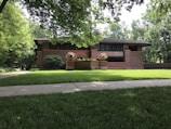 A brick house with a low-pitched roof surrounded by lush greenery and a well-maintained lawn. There are vibrant flowers planted in large planters near the entrance, and the architecture features horizontal lines and large windows.