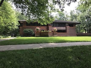 A brick house with a low-pitched roof surrounded by lush greenery and a well-maintained lawn. There are vibrant flowers planted in large planters near the entrance, and the architecture features horizontal lines and large windows.