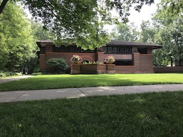 A brick house with a low-pitched roof surrounded by lush greenery and a well-maintained lawn. There are vibrant flowers planted in large planters near the entrance, and the architecture features horizontal lines and large windows.