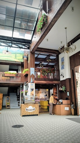 A spacious indoor area with wooden beams and a high glass ceiling. There are decorative plants and a Bavarian theme with flags and a mural of a pastoral landscape. A person stands at a wooden counter, possibly making a purchase. The area includes a food display case and several pieces of rustic furniture.