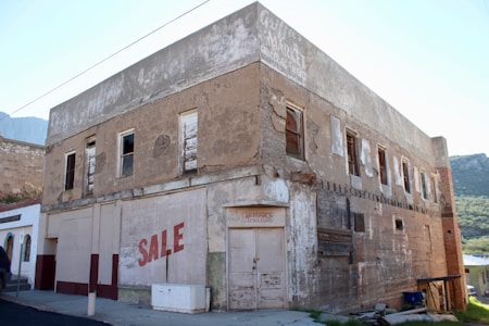 An old, weathered, two-story building with peeling paint and boarded-up windows. The facade has signs of neglect, with a prominent 'SALE' sign painted on it. The structure seems to have been a market or general store in the past, as indicated by faded signage at the top. The area around the building is quiet, with a mountainous landscape visible in the background.