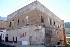 An old, weathered, two-story building with peeling paint and boarded-up windows. The facade has signs of neglect, with a prominent 'SALE' sign painted on it. The structure seems to have been a market or general store in the past, as indicated by faded signage at the top. The area around the building is quiet, with a mountainous landscape visible in the background.