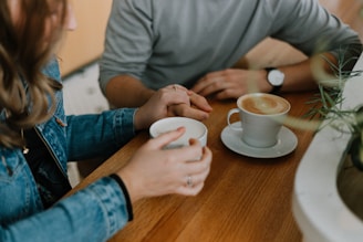 Two people are sitting at a wooden table, each holding a cup of coffee. One hand is gently holding the other person's hand, suggesting a moment of connection. A watch is visible on one person's wrist. The setting includes casual attire and a cozy atmosphere with soft natural lighting.