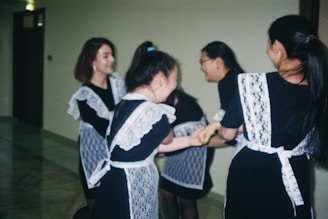 A group of young women wearing dark dresses with white lace aprons are in a hallway, seemingly engaging in a playful or joyous interaction, possibly dancing or spinning together while smiling.