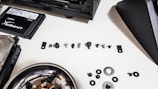 A collection of various small screws, washers, and computer components are arranged on a white surface. To the left, there is a solid-state drive labeled 'HyperX Fury' and a hard disk drive. A metallic bowl containing a few more screws and washers is also present. Parts of a computer case can be seen at the top and center right of the image.