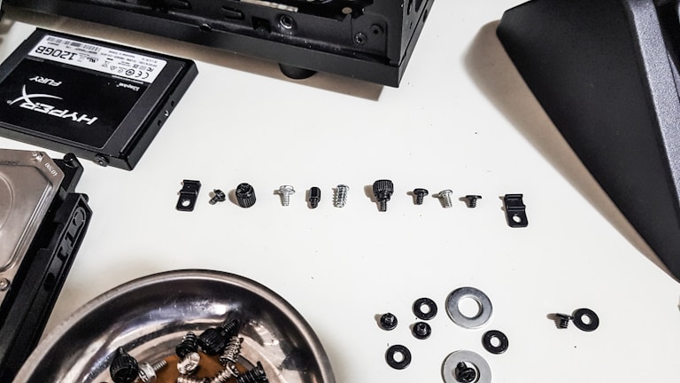 A collection of various small screws, washers, and computer components are arranged on a white surface. To the left, there is a solid-state drive labeled 'HyperX Fury' and a hard disk drive. A metallic bowl containing a few more screws and washers is also present. Parts of a computer case can be seen at the top and center right of the image.