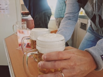 Several people gather around a table, with one individual handing over a glass mug filled with frothy beer. The setting appears casual, possibly at a bar or a social gathering. Background elements include another person standing by a counter and a menu or price list on the wall.