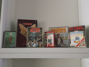 Several retro video game boxes are displayed on a shelf. The collection includes titles such as World Cup Football, Star Wars, Impossible Mission II, Tetris, and Ghostbusters. The games appear to be for older gaming systems, indicated by the distinctive box designs and logos.