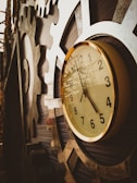 A detailed close-up of a vintage clock with a gold-rimmed frame is mounted on an industrial, gear-like structure. The clock face shows the time as ten past ten, and the gears around it create a steampunk aesthetic.