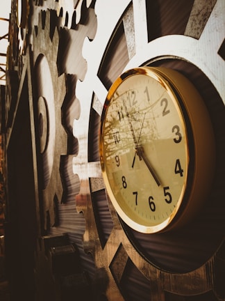 A detailed close-up of a vintage clock with a gold-rimmed frame is mounted on an industrial, gear-like structure. The clock face shows the time as ten past ten, and the gears around it create a steampunk aesthetic.