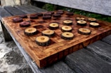 A rustic chessboard made from dark and light wooden slices set on a checkered wooden board. The pieces resemble cross-sections of tree branches, enhancing the natural aesthetic. The board rests on an outdoor wooden bench, providing a serene and earthy setting.