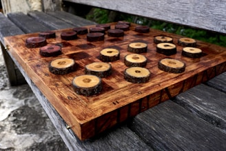 A rustic chessboard made from dark and light wooden slices set on a checkered wooden board. The pieces resemble cross-sections of tree branches, enhancing the natural aesthetic. The board rests on an outdoor wooden bench, providing a serene and earthy setting.