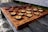 A rustic chessboard made from dark and light wooden slices set on a checkered wooden board. The pieces resemble cross-sections of tree branches, enhancing the natural aesthetic. The board rests on an outdoor wooden bench, providing a serene and earthy setting.