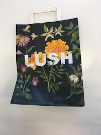 A paper bag with floral designs, including vibrant orange and pink flowers, green leaves, and bees, featuring the word 'LUSH' in large white letters.