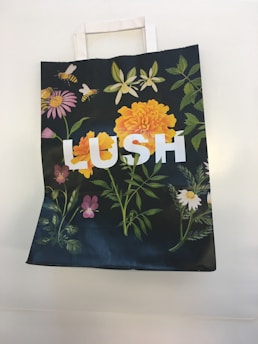 A paper bag with floral designs, including vibrant orange and pink flowers, green leaves, and bees, featuring the word 'LUSH' in large white letters.