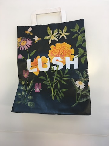 A paper bag with floral designs, including vibrant orange and pink flowers, green leaves, and bees, featuring the word 'LUSH' in large white letters.