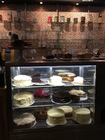 A display case filled with a variety of cakes in a café setting. The top shelf contains different types of cakes, including chocolate and vanilla flavors. A glass shelf behind the case holds cups, glasses, and decorative cans against a mosaic tile wall.