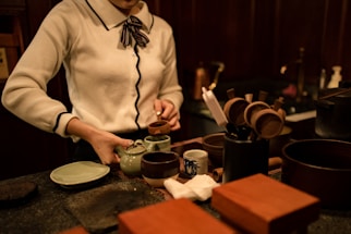 A person is preparing tea in a traditional setting, with a teapot, cups, and various utensils arranged on a wooden counter. The person is dressed in a white sweater with a ribbon tie at the collar and is pouring tea into a small cup.