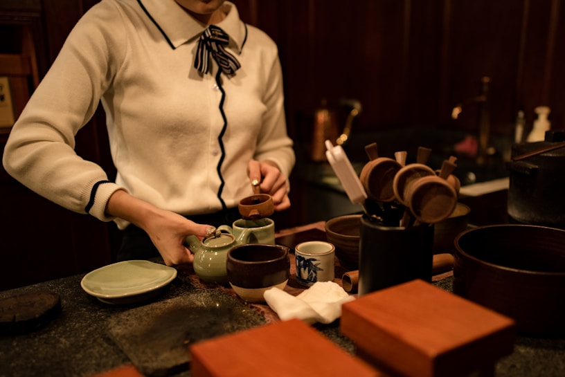 A person is preparing tea in a traditional setting, with a teapot, cups, and various utensils arranged on a wooden counter. The person is dressed in a white sweater with a ribbon tie at the collar and is pouring tea into a small cup.