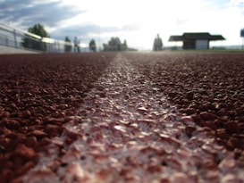 A close-up view of an outdoor track surface, with a white line running down the center. The focus is on the texture of the track material, with the background showing a blurred image of people and structures against a bright sky.