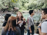 A group of young people stands by a clear turquoise spring surrounded by lush greenery. The individuals appear engaged in conversation, with some looking at the camera while others face away. The background features wooden cottages and dense forest, evoking a natural and serene environment.