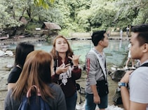 A group of young people stands by a clear turquoise spring surrounded by lush greenery. The individuals appear engaged in conversation, with some looking at the camera while others face away. The background features wooden cottages and dense forest, evoking a natural and serene environment.