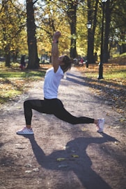 A person is performing a lunge exercise on a dirt path in a park surrounded by tall trees. The sunlight filters through the leaves, casting dappled shadows on the ground. The person is dressed in a white shirt and black leggings, with light pink athletic shoes. The autumn foliage has started to turn yellow and brown, creating a warm, serene atmosphere.