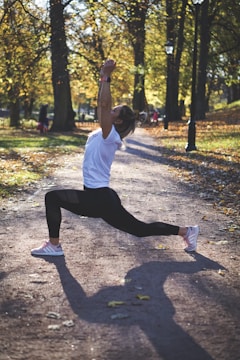 A person is performing a lunge exercise on a dirt path in a park surrounded by tall trees. The sunlight filters through the leaves, casting dappled shadows on the ground. The person is dressed in a white shirt and black leggings, with light pink athletic shoes. The autumn foliage has started to turn yellow and brown, creating a warm, serene atmosphere.