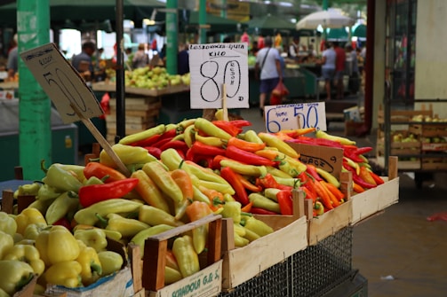 A vibrant market scene with a variety of peppers in crates prominently displayed at the foreground. The peppers are in shades of red, yellow, and green. Pricing signs are visible, written in a foreign language, indicating the cost of the produce. The background shows a bustling atmosphere with blurred people and other market stalls under green canopies.