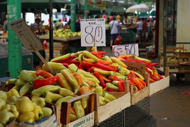 A vibrant market scene with a variety of peppers in crates prominently displayed at the foreground. The peppers are in shades of red, yellow, and green. Pricing signs are visible, written in a foreign language, indicating the cost of the produce. The background shows a bustling atmosphere with blurred people and other market stalls under green canopies.