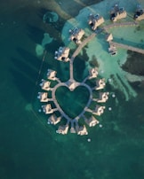 Aerial view of overwater bungalows arranged in a heart shape surrounded by clear turquoise waters. The structures are connected by wooden pathways, and small white circular floats or buoys are visible in the water.