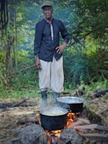 A man is standing in a lush, green forest clearing beside a campfire. Two black pots are placed on the fire, surrounded by rocks and logs. He is dressed in a black shirt, light-colored pants, and boots, with a cap on his head. Smoke rises from the boiling pots.