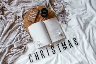 A cozy setup includes an open book on a white surface with the word 'CHRISTMAS' printed in bold black letters. Nearby, a round wooden tray holds a Canon camera lens, a glittery phone case with the phrase 'THE FUTURE IS FEMALE,' and a textured beige blanket. String lights with star-shaped bulbs add a warm, festive touch.