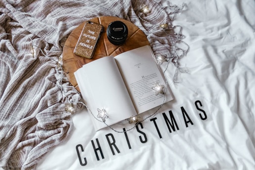 A cozy setup includes an open book on a white surface with the word 'CHRISTMAS' printed in bold black letters. Nearby, a round wooden tray holds a Canon camera lens, a glittery phone case with the phrase 'THE FUTURE IS FEMALE,' and a textured beige blanket. String lights with star-shaped bulbs add a warm, festive touch.