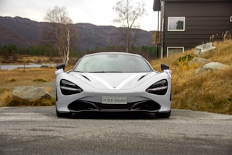 A sleek, high-performance sports car is parked on a paved road surrounded by a rural landscape. The car is metallic silver with aerodynamic lines and black accents. In the background, there is a wooden house on the right, rocky landscape, dry grasses, and barren trees under an overcast sky.