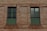 A brick wall with two adjacent windows featuring green shutters and black metal bars running vertically and horizontally.