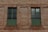 A brick wall with two adjacent windows featuring green shutters and black metal bars running vertically and horizontally.