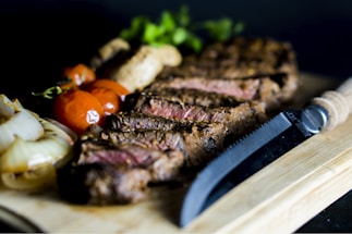 A perfectly grilled steak sliced into pieces resting on a wooden board, accompanied by roasted cherry tomatoes, mushrooms, and grilled onions. A knife lies nearby for serving.