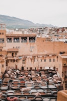 An ancient tannery with numerous circular dyeing pits occupies the foreground. Workers are seen handling hides, surrounded by large stone vessels containing various colored dyes. The buildings in the background are constructed with earthy tones, typical of a historic city, and there are people watching from the balconies. In the distance, a mountain range completes the scenic backdrop.
