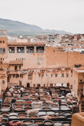 An ancient tannery with numerous circular dyeing pits occupies the foreground. Workers are seen handling hides, surrounded by large stone vessels containing various colored dyes. The buildings in the background are constructed with earthy tones, typical of a historic city, and there are people watching from the balconies. In the distance, a mountain range completes the scenic backdrop.